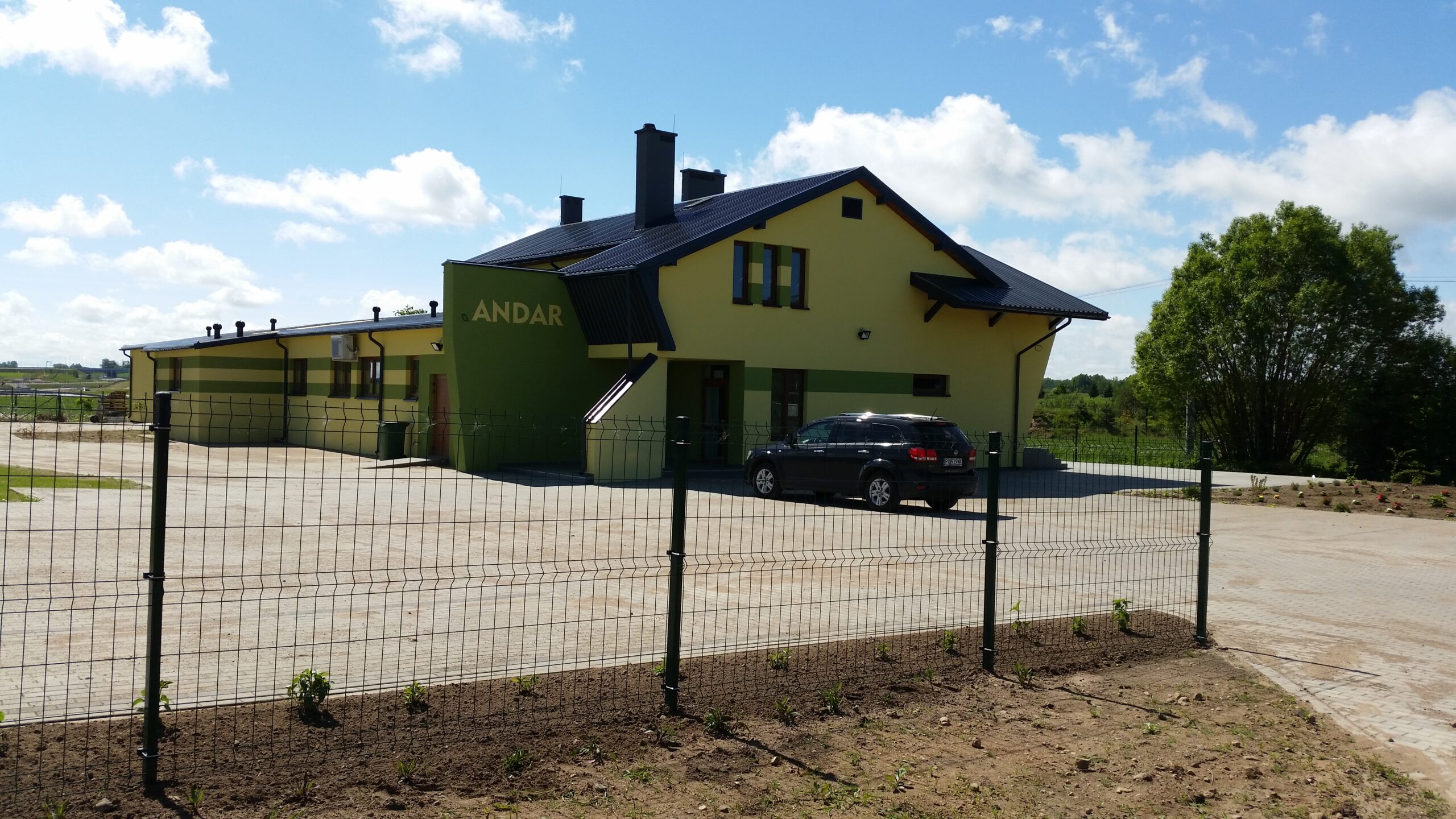 Żółto-zielony budynek firmy ANDAR z parkingiem i czarnym samochodem.